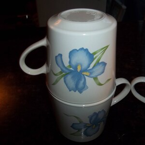 May include: Two white ceramic mugs with blue and green iris flower designs. The mugs are stacked on top of each other.