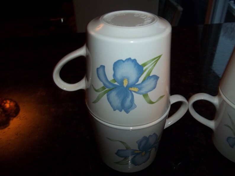 May include: Two white ceramic mugs with blue and green iris flower designs. The mugs are stacked on top of each other.