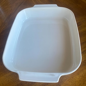 May include: A rectangular, off-white baking dish with rounded corners and two handles. The dish is made of a smooth, glossy material and sits on a wooden surface. This versatile dish is suitable for baking and serving.