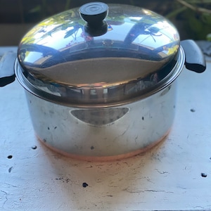 May include: A stainless steel pot with a lid and black handles. The pot has a shiny, reflective surface and a copper-colored band at the base. The lid has a black knob. The pot is sitting on a white surface.