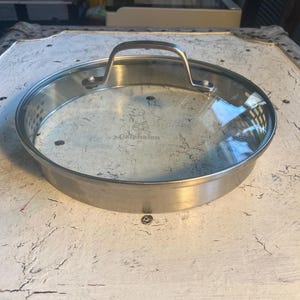 May include: A stainless steel Calphalon pan lid with a clear glass center and a metal handle. The lid is round and has a silver finish. The Calphalon logo is visible on the glass. The lid is designed for cookware.