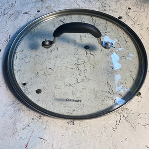 May include: A clear glass Cuisinart pot lid with a black handle and a stainless steel rim. The lid is round and has a black handle. The Cuisinart logo is visible on the glass.