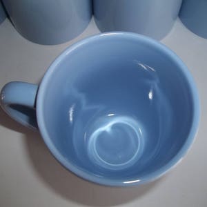 May include: A light blue ceramic mug with a handle, viewed from above. The mug has a smooth, glossy finish and a rounded interior. Several other mugs are visible in the background. The mugs are likely for beverages.