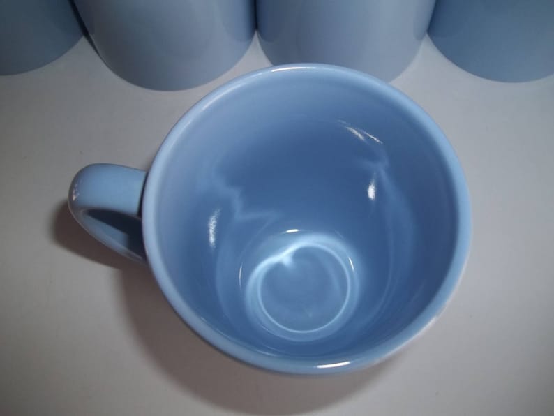 May include: A light blue ceramic mug with a handle, viewed from above. The mug has a smooth, glossy finish and a rounded interior. Several other mugs are visible in the background. The mugs are likely for beverages.