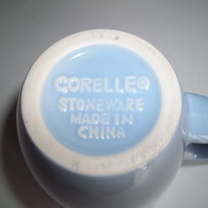 May include: A light blue Corelle stoneware mug, viewed from the bottom. The base is light blue with a white rim. The text "CORELLEO STONEWARE MADE IN CHINA" is embossed in white.