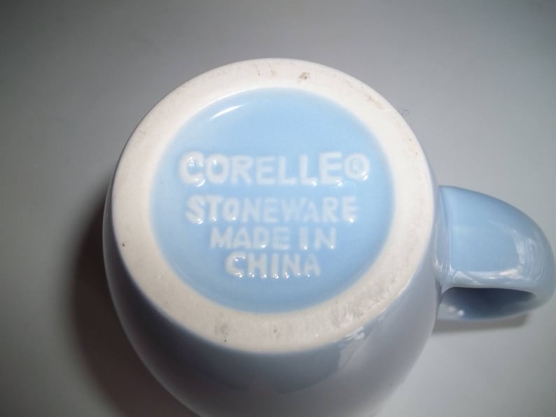 May include: A light blue Corelle stoneware mug, viewed from the bottom. The base is light blue with a white rim. The text "CORELLEO STONEWARE MADE IN CHINA" is embossed in white.