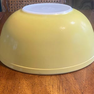 May include: A yellow mixing bowl with a white interior rim. The bowl is made of a glossy material and sits on a wooden surface. The bowl is round and has a classic design, suitable for kitchen use.