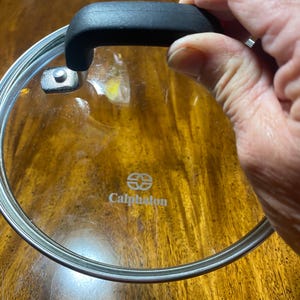 May include: A clear glass lid with a black handle and a silver rim. The lid has the Calphalon logo printed on it.