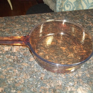 May include: A clear, amber-colored glass skillet with a long handle. The skillet is round with a slightly flared rim. The handle is an extension of the skillet's body. The skillet is sitting on a speckled brown countertop.
