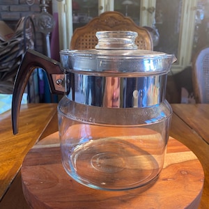 May include: A clear glass coffee pot with a stainless steel band and a dark brown handle. The pot sits on a round wooden serving tray. The lid is also glass. The background includes a wooden table and a window.
