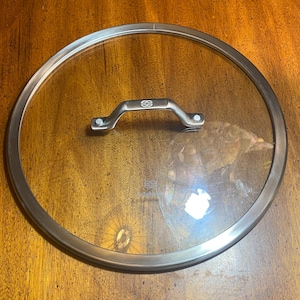May include: A clear glass pot lid with a stainless steel rim and handle. The handle is curved and attached to the lid with two rivets. The lid is circular and designed to fit on a pot or pan.