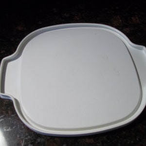 May include: A white, square-shaped plastic lid with rounded corners and integrated handles. The lid has a slightly textured surface and is designed to fit a container. The handles are curved and provide a secure grip.