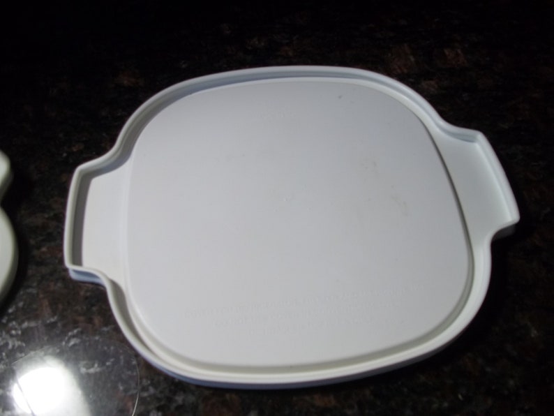 May include: A white, square-shaped plastic lid with rounded corners and integrated handles. The lid has a slightly textured surface and is designed to fit a container. The handles are curved and provide a secure grip.