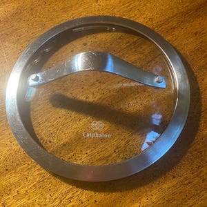 May include: A clear glass Calphalon brand pot lid with a stainless steel rim and handle. The lid is circular and sits on a wooden surface. The handle is curved and attached to the glass with rivets.