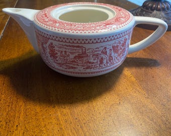 May include: A white ceramic teapot with a red transferware design. The teapot features a spout, handle, and a lid opening. The red design depicts a pastoral scene with figures and buildings. The teapot is sitting on a wooden surface.