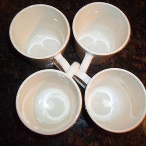 May include: Four white ceramic mugs with rounded handles. The mugs are arranged in a circle with the handles pointing outwards.