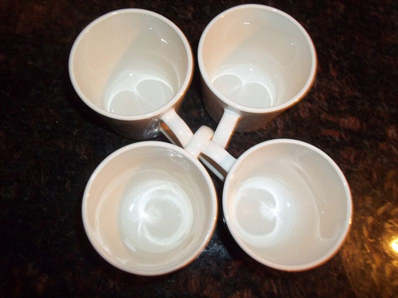 May include: Four white ceramic mugs with rounded handles. The mugs are arranged in a circle with the handles pointing outwards.