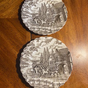 May include: Two round ceramic plates with scalloped edges, featuring a monochromatic gray and white transferware scene of a horse-drawn carriage arriving at a timber-framed inn with tall trees, rolling hills, and a cloudy sky in the background.