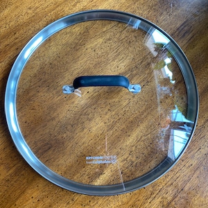May include: A clear glass Kitchen Essentials from Calphalon lid with a stainless steel rim and a black handle. The lid is circular and designed for cookware.