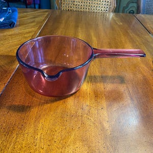 May include: A vintage, reddish-brown glass saucepan with a long handle. The saucepan has a pouring spout and a rounded bowl shape. It sits on a wooden table, showcasing its classic design and functionality. This kitchenware item is ideal for cooking and serving.