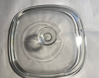 May include: A clear, square glass lid with rounded corners. The lid has a small, circular handle in the center and is designed to fit a square pot or pan. The glass is transparent, allowing visibility of the contents.