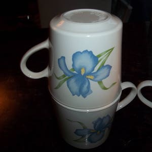 May include: Two white ceramic mugs with blue and green iris flower designs. The mugs are stacked on top of each other.
