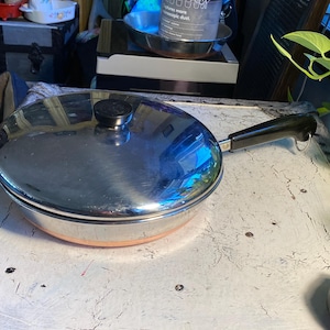 May include: A stainless steel frying pan with a lid and a black handle. The pan has a copper-colored rim and a black knob on the lid. The pan is sitting on a white surface.