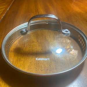 May include: A clear glass Cuisinart lid with a stainless steel rim and handle. The lid has a circular shape with small ventilation holes around the edge. The handle is arched and attached with rivets.
