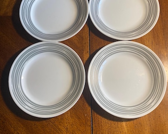 May include: A set of four round white plates, each featuring a series of concentric gray circular lines around the rim. The plates have a smooth, glossy finish and a clean, circular design, providing a uniform appearance across all four pieces.
