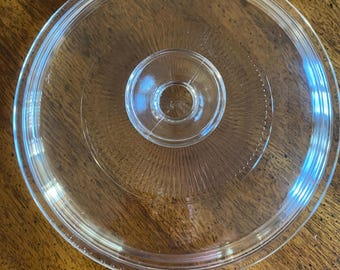 May include: A clear glass lid with a circular design. The lid has a raised, circular handle in the center and a slightly curved edge. The lid is designed for cookware and is made of transparent glass.