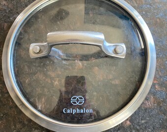 May include: A round, clear glass pot lid with a brushed metal rim and a matching brushed metal handle. The lid features the white Calphalon logo printed on the glass surface. The handle is secured to the lid with two visible metal rivets.