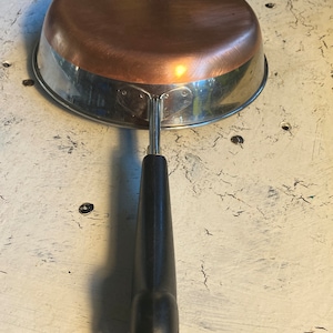 May include: A copper-bottomed frying pan with a stainless steel rim and a black handle. The pan is round and has a shiny copper interior. The handle is black and has a loop for hanging.