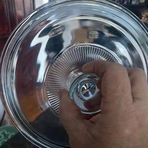 May include: Clear glass lid with a fluted edge and a knob on top. The lid is designed to fit on a glass dish or bowl.