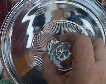 May include: Clear glass lid with a fluted edge and a knob on top. The lid is designed to fit on a glass dish or bowl.