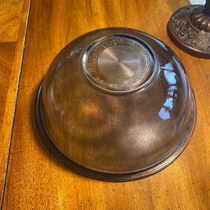 May include: A clear, brown glass mixing bowl with a circular base and a wide rim. The bowl is sitting on a wooden surface. The bottom of the bowl has a circular design with text.