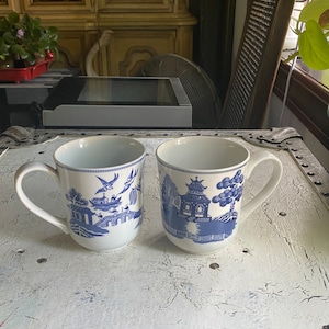 May include: Two white ceramic mugs with blue and white floral designs. The mugs have a handle on the right side and are sitting on a white wooden surface.