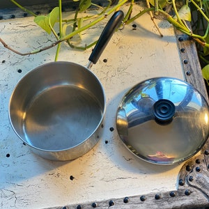 May include: A stainless steel saucepan with a black handle and a matching lid. The pan is round and has a shiny, reflective surface. The lid has a black knob. The pan is sitting on a white surface.