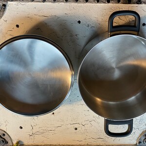 May include: Two stainless steel pots with black handles. The pots are empty and sitting on a white surface.