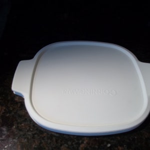 May include: A white CorningWare lid with a square shape and rounded corners. The lid has two handles on opposite sides and the brand name is embossed in the center. The lid is designed for cookware.