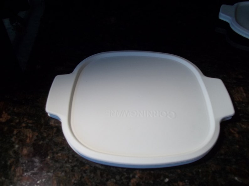 May include: A white CorningWare lid with a square shape and rounded corners. The lid has two handles on opposite sides and the brand name is embossed in the center. The lid is designed for cookware.