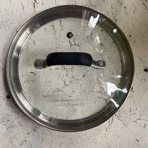 May include: A silver and glass lid with a black handle. The lid has the text "Kitchen Essentials from Calphalon" printed on it.
