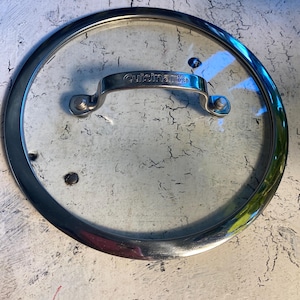 May include: A clear glass Cuisinart lid with a stainless steel rim and handle. The handle is curved and attached to the glass with two rivets. The lid is circular and designed for cookware.