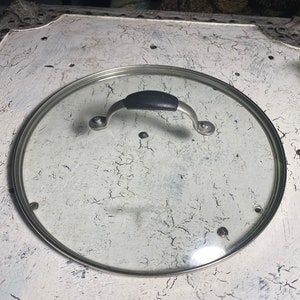 May include: A clear glass pot lid with a stainless steel rim and a black handle. The lid has a silver handle and is designed to fit a pot. The lid is round and has a clear glass surface.