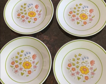 May include: Four white ceramic plates with a floral design. Each plate features a yellow flower at the center, surrounded by smaller orange, pink, and blue flowers. A green border encircles the rim of each plate, creating a cohesive set.
