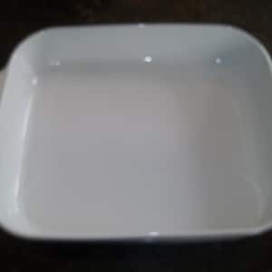May include: A white, rectangular baking dish with rounded corners and two handles. The dish is empty and appears to be made of ceramic or a similar material. The background is a dark, solid color.