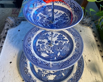May include: A three-tiered serving stand with blue and white Willow pattern plates. The plates are stacked with a metal handle and a red ribbon. The stand is on a distressed white surface.
