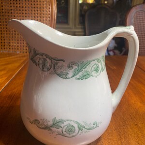 May include: White ceramic pitcher with a spout and handle. The pitcher is decorated with green floral and scrollwork patterns around the body. The pitcher is sitting on a wooden table.