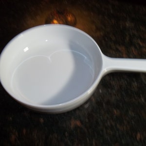 May include: A white ceramic pan with a long handle. The pan is round with a heart-shaped indentation on the bottom. The handle is straight and extends from the side of the pan. The pan is sitting on a dark, speckled surface.