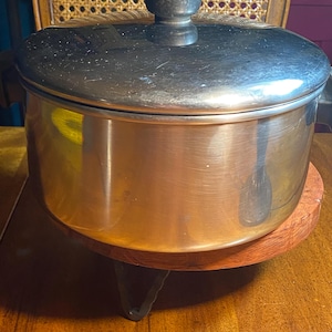 May include: A copper-colored cooking pot with a dark metal lid and a black handle. The pot sits on a round, wooden trivet with three black metal legs. The pot is on a wooden table.