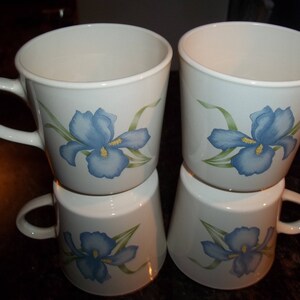 May include: Four white ceramic mugs with blue and green iris flower designs. The mugs are stacked in pairs.